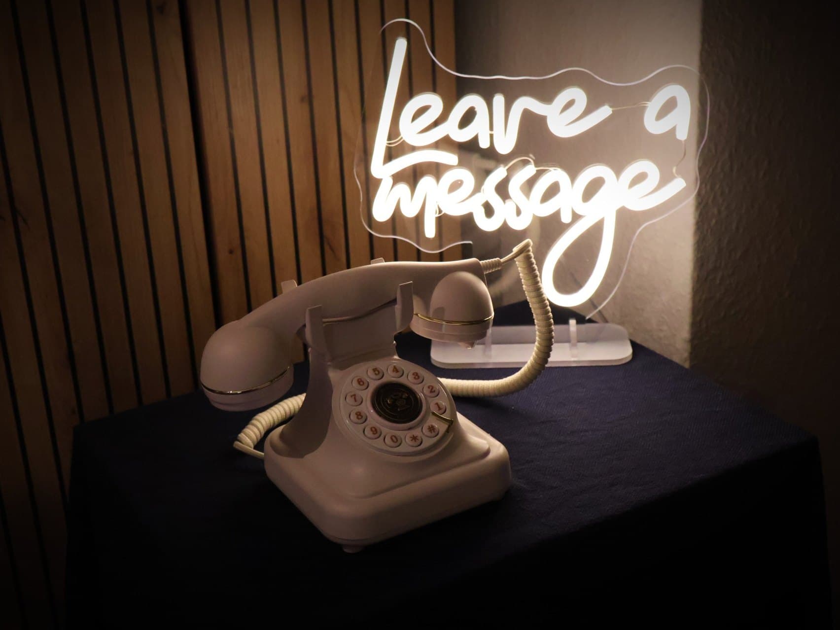 Cremefarbenes Retro-Telefon des Audiogästebuchs auf einem dunkel eingedeckten Tisch, dahinter ein leuchtendes Neon-Schild mit der Aufschrift „Leave a message“.